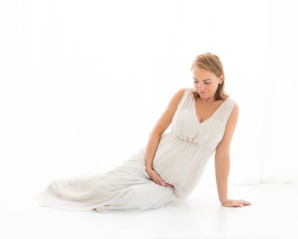 Taunton-Photographer-Sharon-Lewis-pregnancy-photos Pregnant lady if white dress sitting on floor holding her bump