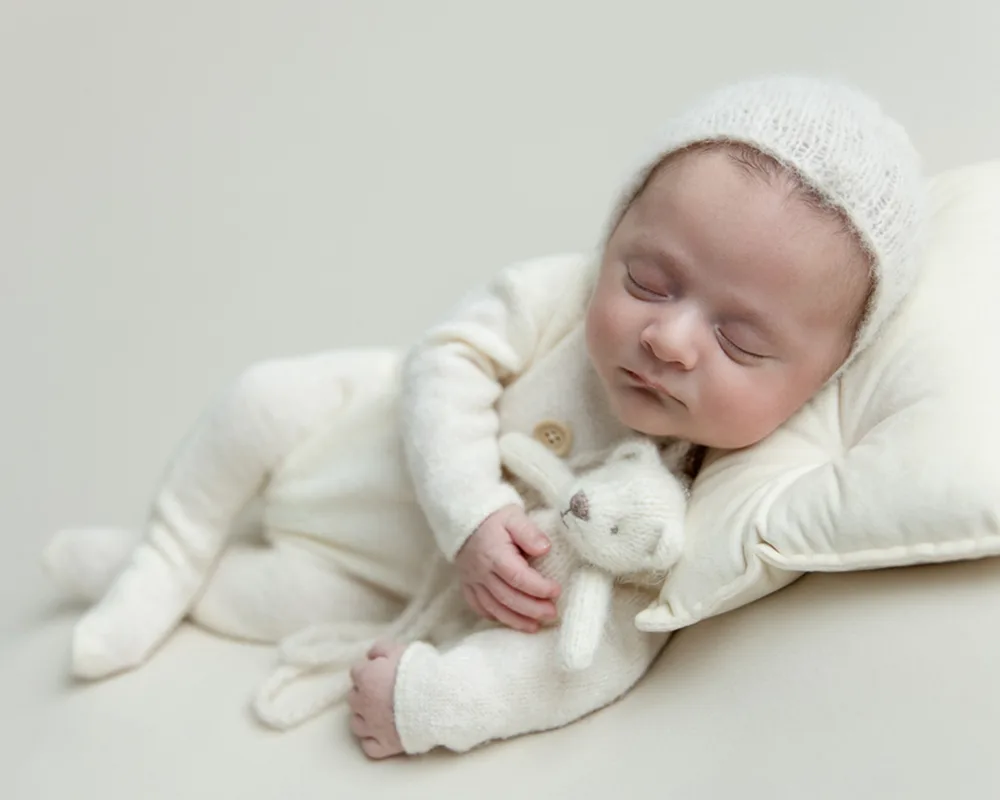 Baby boy in cream outfit and bonnet snuggling a teddy