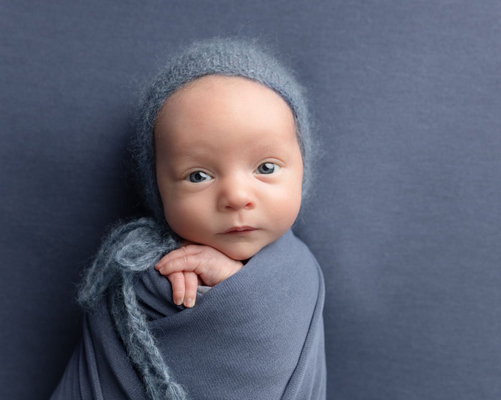 Baby boy wrapped in blue with a blue bonnet looking at the camera